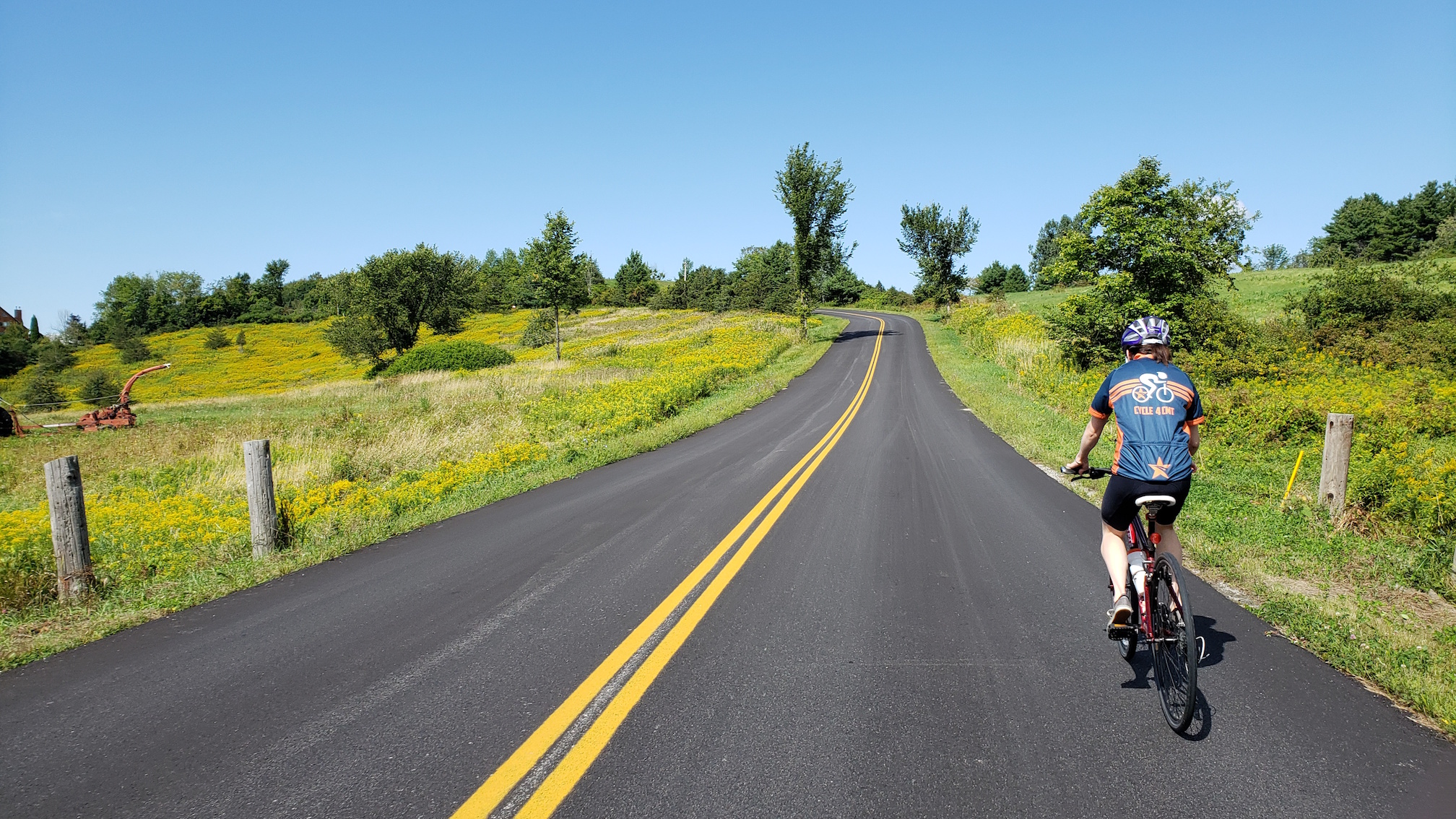 Mary Cate, a woman wearing a helmet and a blue shirt that says “Cycle 4 CMT”, rides her bicycle down a long straight road which cuts through a beautiful lush landscape of grass and sparse trees.